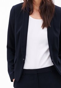 Námořnický blazer s klopovými límečky, zapínáním na jeden knoflík, dvěma předními kapsami a hladkým materiálem. Nositý přes bílou halenku. Přiléhavý střih.