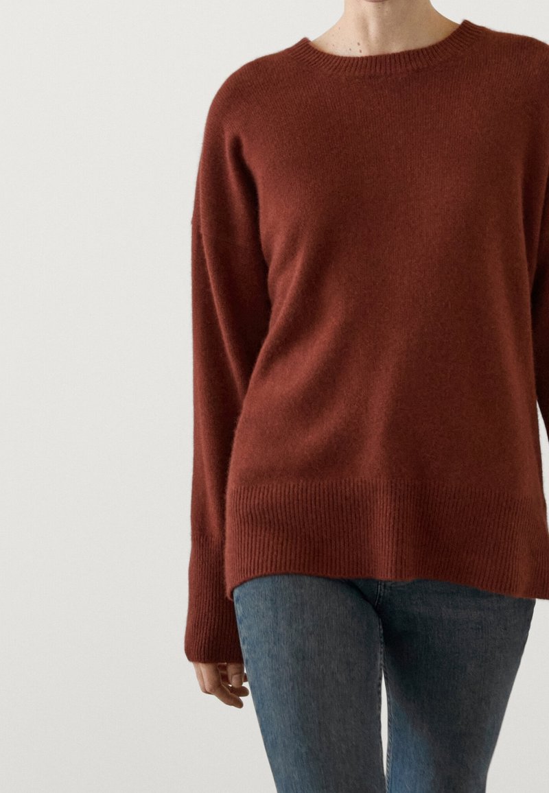 Pull de couleur rouille avec un col rond et des manches longues, doté d'un ourlet et de poignets côtelés, fabriqué à partir d'un matériau doux et texturé.