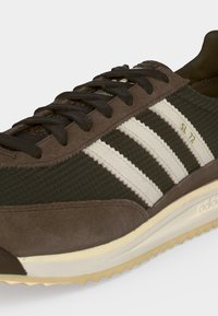 Καφέ και ελαιί Adidas SL 72 αθλητικό παπούτσι με λευκές ρίγες, μαύρα κορδόνια, υφασμάτινη υφή και ανοιχτό μπεζ σόλα με οδοντωτό πέλμα.