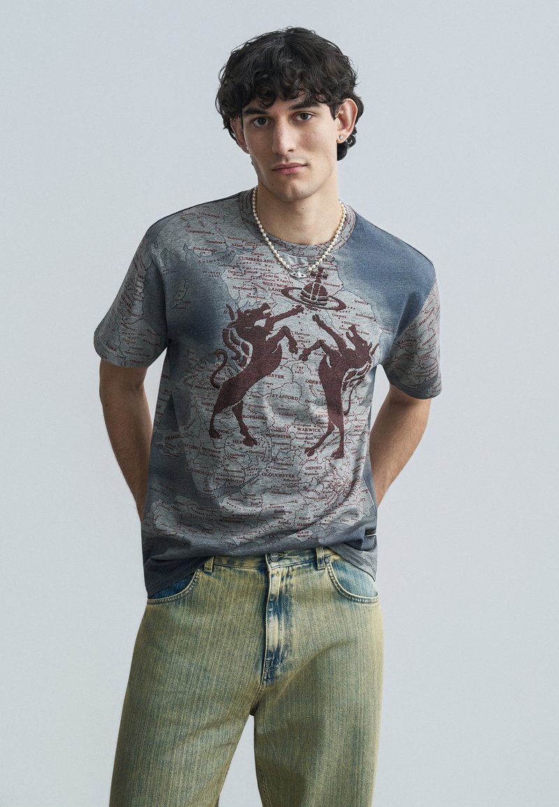 Κοντομάνικο μπλουζάκι με σχέδιο σε γκρι χρώμα, με μια σχεδίαση που μοιάζει με χάρτη και κόκκινες σιλουέτες δύο αλόγων. Τζιν παντελόνια με ξεθωριασμένη κίτρινη απόχρωση.