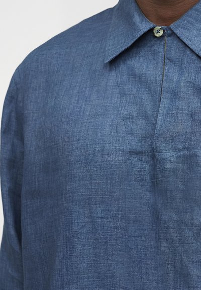 Κοντινό πλάνο ενός ατόμου που φορά ένα μπλε λινό πουκάμισο με μυτερό γιακά και ένα μόνο ορατό κουμπί που είναι κουμπωμένο στο πάνω μέρος.