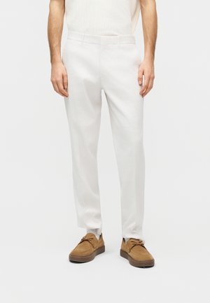 Mies pukeutuneena luonnonvalkoisiin räätälöityihin housuihin, valkoisiin sukkiin ja ruskeisiin mokkanahkaisiin nauhakengiin, seisomassa yksinkertaista vaaleaa taustaa vasten.