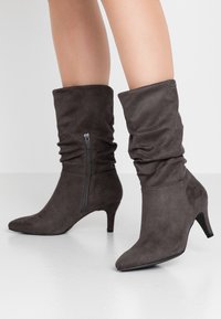 Botas de ante marrón hasta el tobillo con un diseño arrugado, punta afilada, tacón de 5 cm y detalle de cremallera lateral. Textura suave con un ligero brillo.