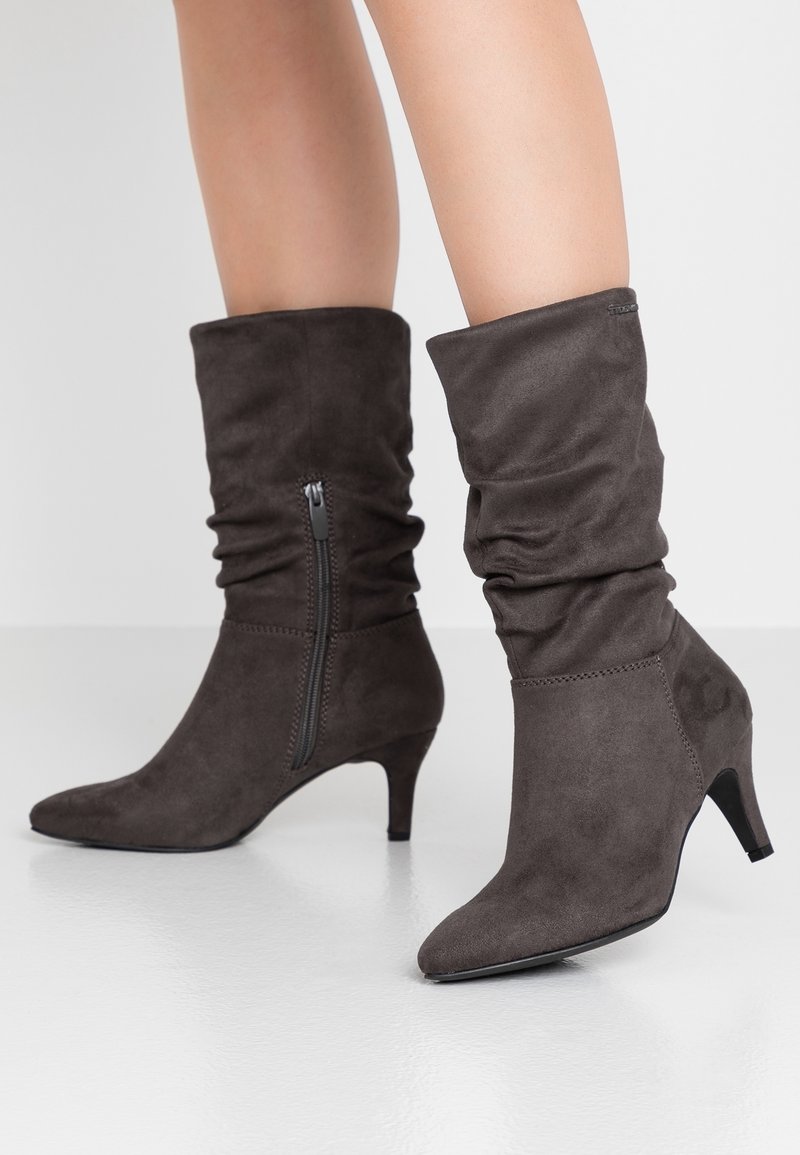 Botas de ante marrón hasta el tobillo con un diseño arrugado, punta afilada, tacón de 5 cm y detalle de cremallera lateral. Textura suave con un ligero brillo.