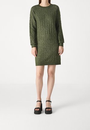 Woman wearing a green knitted long-sleeve dress and black platform sandals, standing against a plain white background.