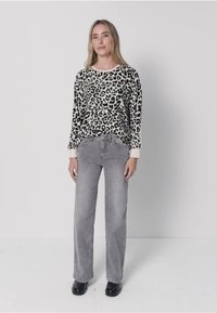 Mustavalkoinen leopardi-kuvioinen neule; harmaat suorat housut; malli seisoo valkoista taustaa vasten; kiiltävät mustat kengät.