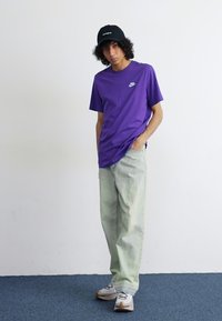 Lilla Nike t-shirt, grønne baggy jeans, sort kasket og beige sneakers. T-shirten har et lille logo på brystet og en afslappet pasform.