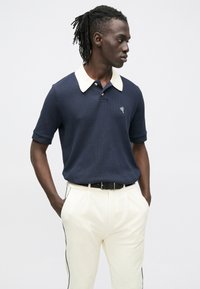 Granatowa koszulka polo z kontrastowym kremowym kołnierzykiem, krótkimi rękawami i małym emblematem w kształcie serca. Połączona z kremowymi spodniami z czarnymi paskami po bokach.