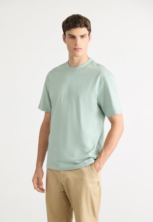 Jeune homme debout, une main dans la poche, portant un t-shirt à manches courtes vert clair et un pantalon beige sur un fond uni.