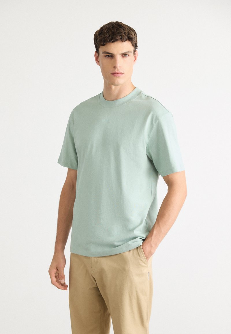 Jovem de pé com uma mão no bolso, vestindo uma t-shirt de manga curta verde claro e calças bejes, contra um fundo simples.
