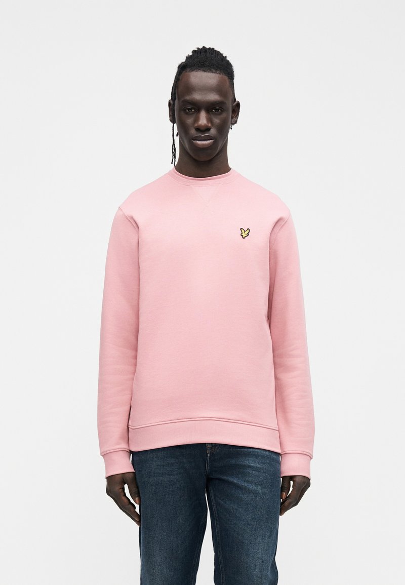 Sweatshirt rosa com gola e punhos canelados, apresenta um pequeno logotipo amarelo de borboleta no peito, combinado com jeans azul escuro.