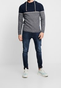 Bluza z kapturem w kolorach granatowym i szarym, wykonana z dzianiny o fakturze, z kontrastującym poziomym paskiem i ozdobami z troczków. Połączona z ciemnymi dżinsami i trampkami.