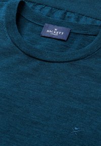 Zbliżenie na sweter w kolorze teal blue z okrągłym dekoltem, z metką Hackett London i haftowanym logo skrzyżowanych lasek blisko brzegu.