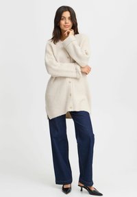 Cardigan beige lavorato a maglia con bottoni, trama a coste, maniche lunghe e spacchi laterali, abbinato a jeans a gamba larga blu scuro e tacchi neri.