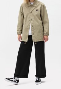 Beige lättviktsjacka med tryckknappar, elastiska ärmmuddar och dragsko i nederkant, tillsammans med svarta vida byxor och svarta sneakers.