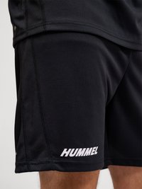 Nærbillede af en person iført sorte træningsshorts med det hvide "HUMMEL"-logo nær kanten på venstre ben.