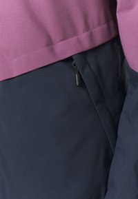Top viola con una texture liscia e sezione inferiore blu scuro, dotato di una tasca laterale con dettaglio zip. Vestibilità regolabile.