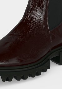 Gros plan d'une botte brillante rouge foncé avec une surface texturée, un panneau latéral élastique et une semelle épaisse noire à crampons profonds.