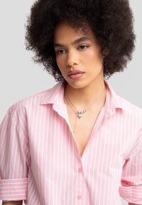 Camisa de botones a rayas rosas y blancas con mangas remangadas, combinada con un collar de plata en capas que presenta un colgante en forma de corazón.