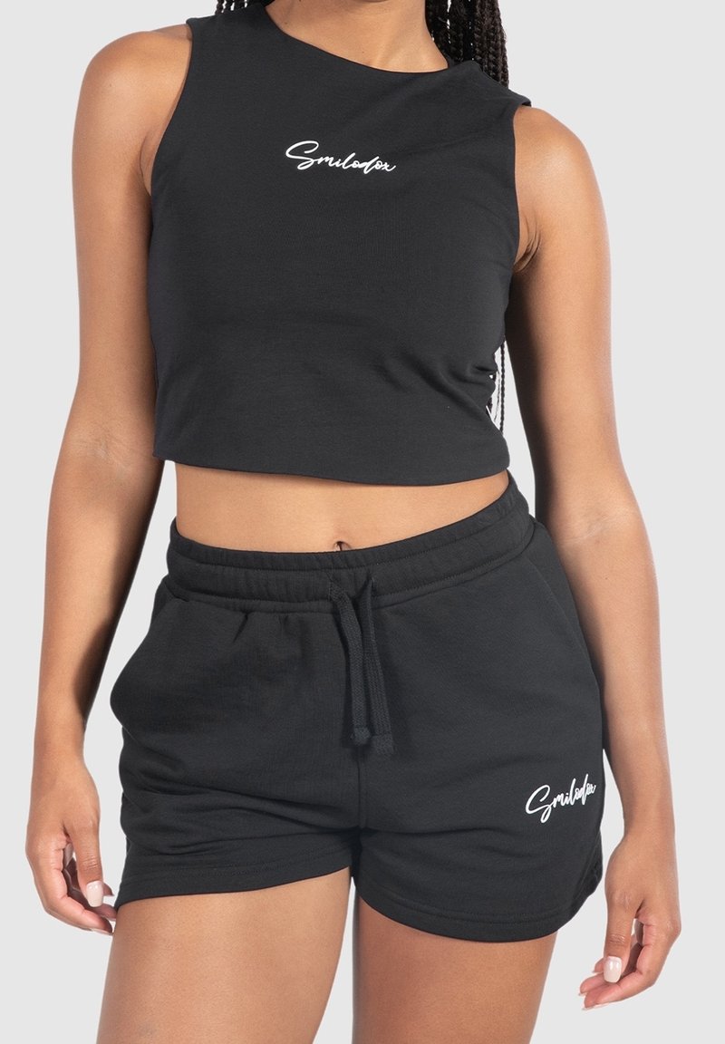 Donna che indossa un crop top senza maniche nero e pantaloni corti neri con coulisse abbinati, entrambi con il logo bianco "Smilodox".
