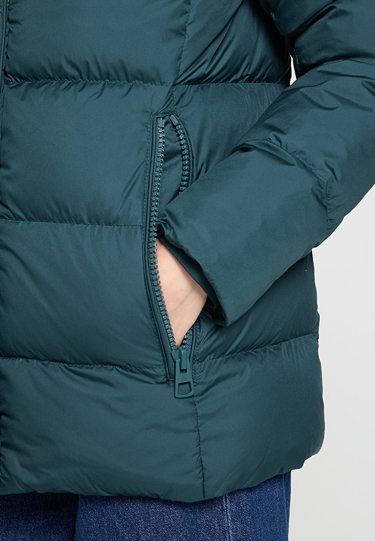 Teal Puffer-Jacke mit sichtbarem Reißverschlussfach, gestepptem Textur und lässiger Passform. Das Material wirkt weich und isoliert.