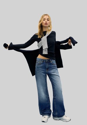 Žena oblečená v tmavom topu s dlhým rukávom, pruhovanom šále, modrých džínsach s širokými nohavicami a bielych teniskách, držiaca námornícku bundu za chrbtom.