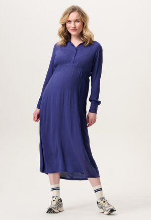 Fließendes, langärmliges, lila Kleid aus leichtem Stoff mit einem Knopfkragen und gerafftem Bund, kombiniert mit Turnschuhen und gestreiften Socken.