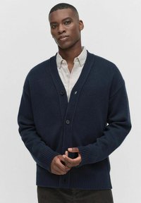 Mörkblå stickad cardigan med v-ringning, långa ärmar, ribbade manschetter och knappstängning framtill, bärs över en ljus skjorta med krage.