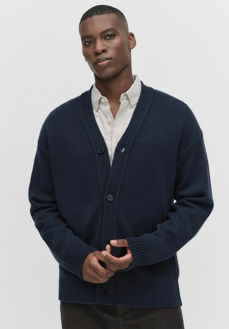 Mörkblå stickad cardigan med v-ringning, långa ärmar, ribbade manschetter och knappstängning framtill, bärs över en ljus skjorta med krage.