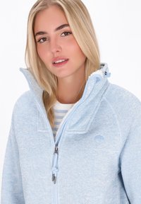 Donna bionda che indossa una giacca in pile leggera blu chiaro con zip e un alto colletto sopra una maglietta a strisce bianca e blu chiaro.
