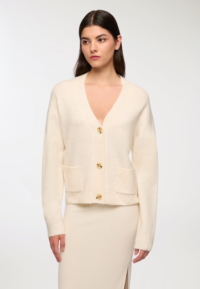 Kremowy kardigan z miękkiego materiału, z głębokim dekoltem w kształcie litery V, czterema dużymi guzikami w złotym kolorze i dwiema przednimi kieszeniami.
