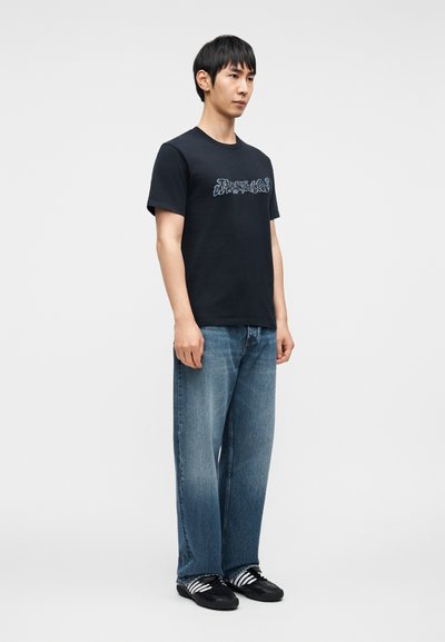 Μαύρο βαμβακερό t-shirt με αντίθετο γραφικό σχέδιο στο μπροστινό μέρος, σε συνδυασμό με ανοιχτό μπλε, φαρδιά τζιν παντελόνια και μαύρα αθλητικά παπούτσια.