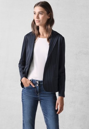Blazer a righe navy con una texture morbida, un bottone singolo e due tasche laterali, abbinato a una maglia bianca e jeans blu.