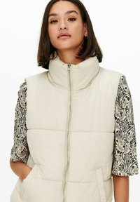 Gilet rembourré en crème clair avec un col haut, fermeture éclair devant et manches courtes noires et beiges à motifs. Comprend des poches latérales.