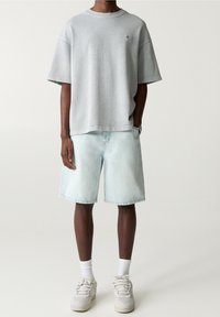 T-shirt oversize in cotone grigio chiaro con maniche corte, abbinata a pantaloni corti in denim blu chiaro. Sneakers neutre e calzini bianchi completano il look.