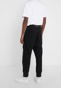 Pantalones de chándal negros con un corte relajado, puños ajustados y costuras anguladas. Presenta un bolsillo trasero y una cinturilla elástica con cordón.