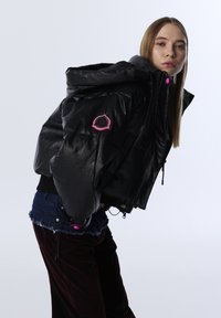 Jonge vrouw die naar voren leunt in een zwarte gewatteerde jas met roze accenten, spijkerbroekshorts en donkere broeken tegen een effen lichte achtergrond.