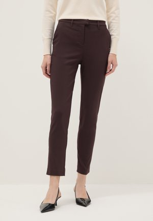 Mujer con pantalones ajustados color marrón oscuro hasta el tobillo, top de manga larga color crema y tacones slingback negros de punta puntiaguda, de pie contra un fondo liso.