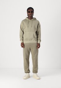 Oliven grøn hoodie og matchende sweatpants, begge lavet af blødt stof. Hoodien har en kanga-pocket; modellen bærer lyse sneakers.