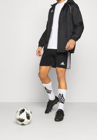 Coupe-vent noir avec capuche, T-shirt blanc, shorts noirs avec rayures blanches, chaussettes blanches avec motif noir, et chaussures de foot noires à côté d'un ballon de football.