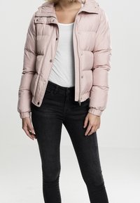 Kvinna som bär en ljusrosa vadderad jacka över en vit tröja och svarta jeans, stående mot en enkel ljus bakgrund.