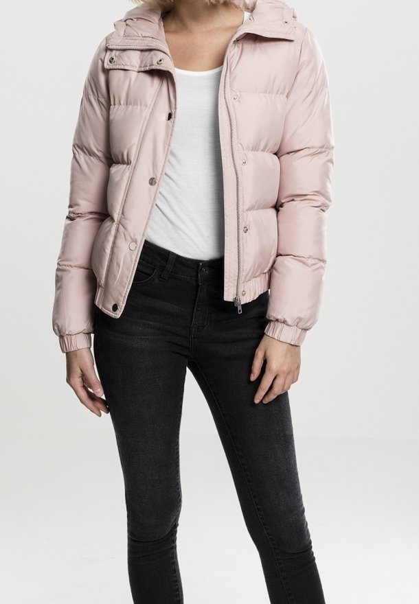 Kvinna som bär en ljusrosa vadderad jacka över en vit tröja och svarta jeans, stående mot en enkel ljus bakgrund.