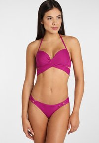 Jasne ružové bikiny s prekríženým bandeau vrchom, s náprsnými popruhmi a výrezmi po bokoch na spodnej časti. Hladká textúra látky.