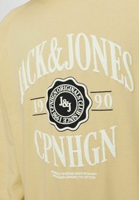 Sweatshirt beige avec texte blanc et un logo circulaire noir. Présente "JACK & JONES," "CPNHGN," et l'année d'établissement 1990 en impression graphique.