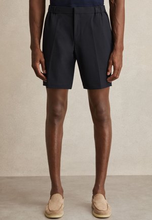 Uomo che indossa pantaloncini neri su misura e scarpe beige senza lacci, in piedi contro uno sfondo chiaro e uniforme.