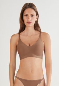 Kvinna som bär en sömlös brun behå och matchande underkläder, står mot en enkel ljus bakgrund, vänd mot kameran.