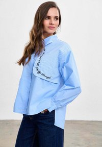 Femeie cu păr lung și castaniu, purtând o cămașă largă bleu deschis cu clapetă cu dungi și inscripție neagră pe piept, asortată cu blugi bleumarin.