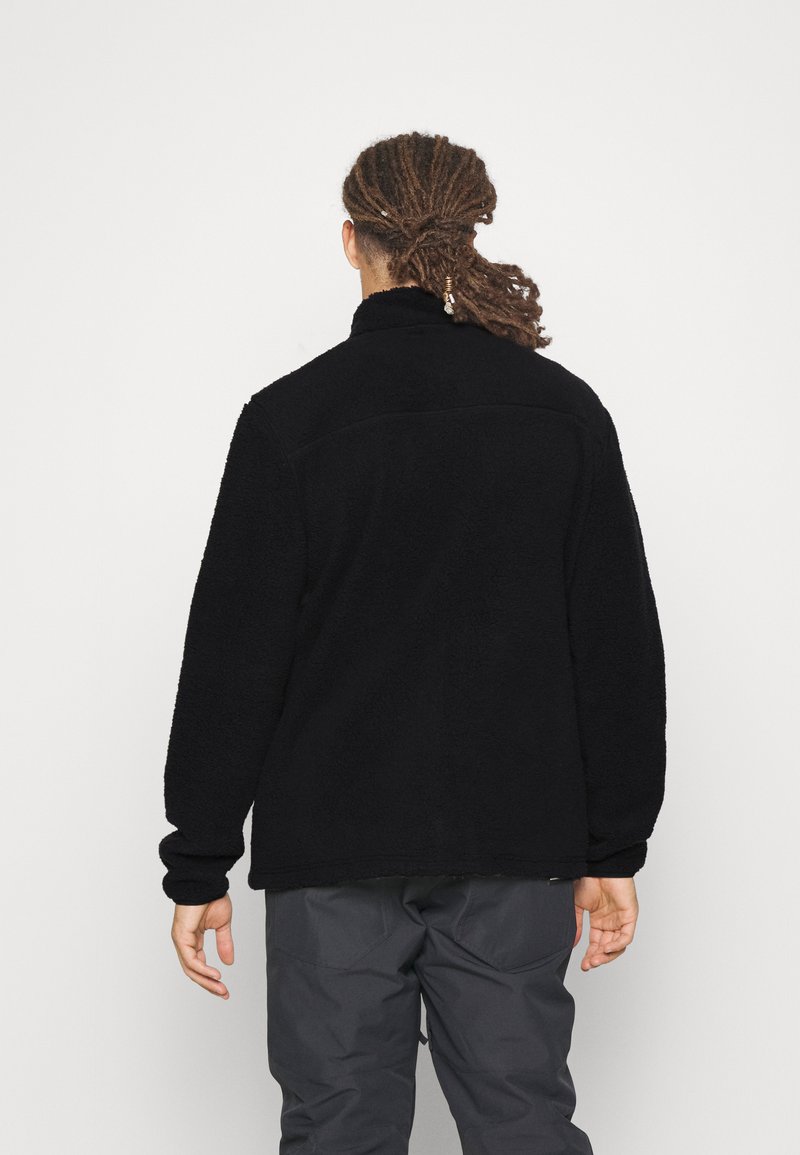 Chaqueta de forro polar negra con un cuello alto y una superficie texturizada. Los puños son ligeramente elásticos. Se lleva con pantalones grises, vista desde la parte de atrás.