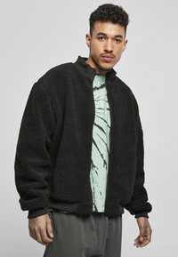 Schwarze Fleecejacke mit Reißverschluss vorne, hohem Kragen und gerippten Bündchen. Strukturiertes, weiches Material; über einem hellgrünen Hemd mit dunklen Mustern getragen.