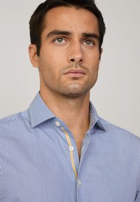 Camicia a strisce blu e bianche con colletto button-down, dotata di un accento giallo lungo la patta dei bottoni. Tessuto morbido, design aderente.